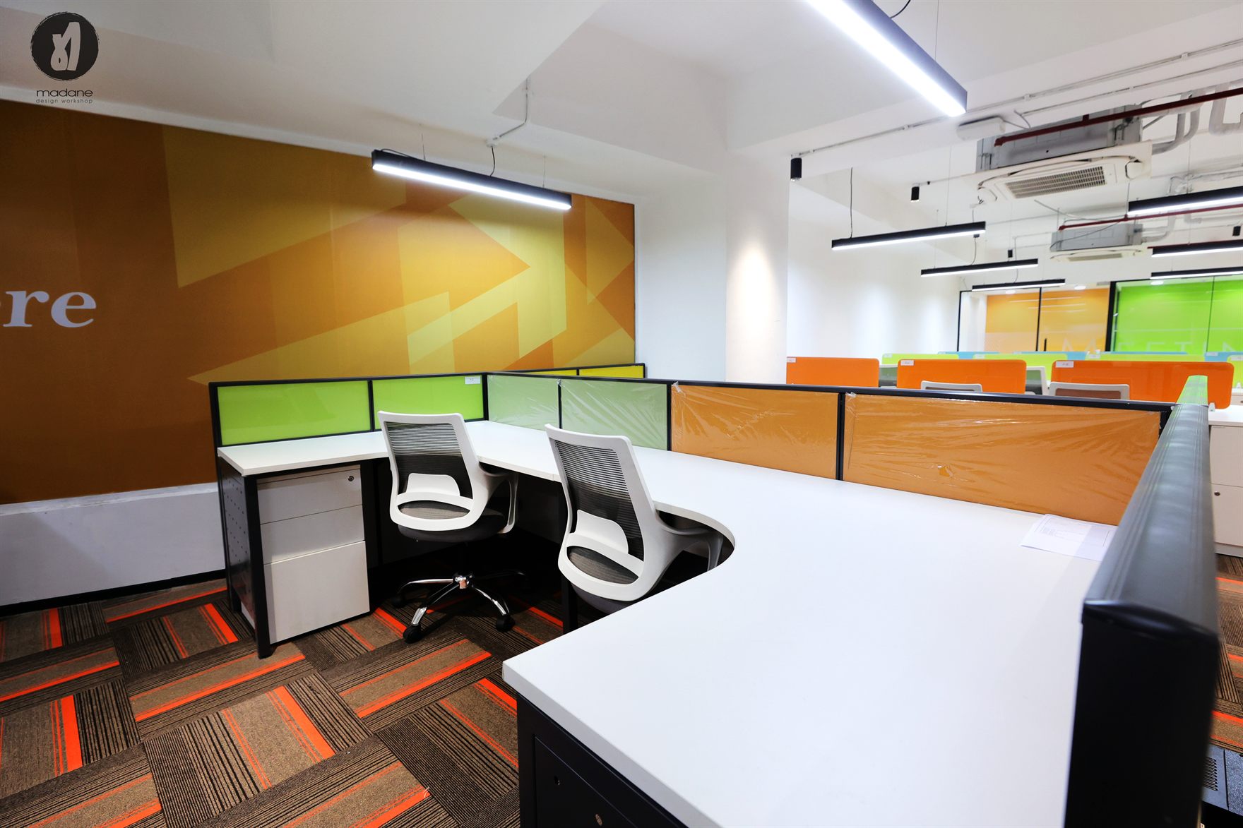 Workstation cluster with green and orange desk screens against a bold ochre arrow graphic wall on striped carpet tiles