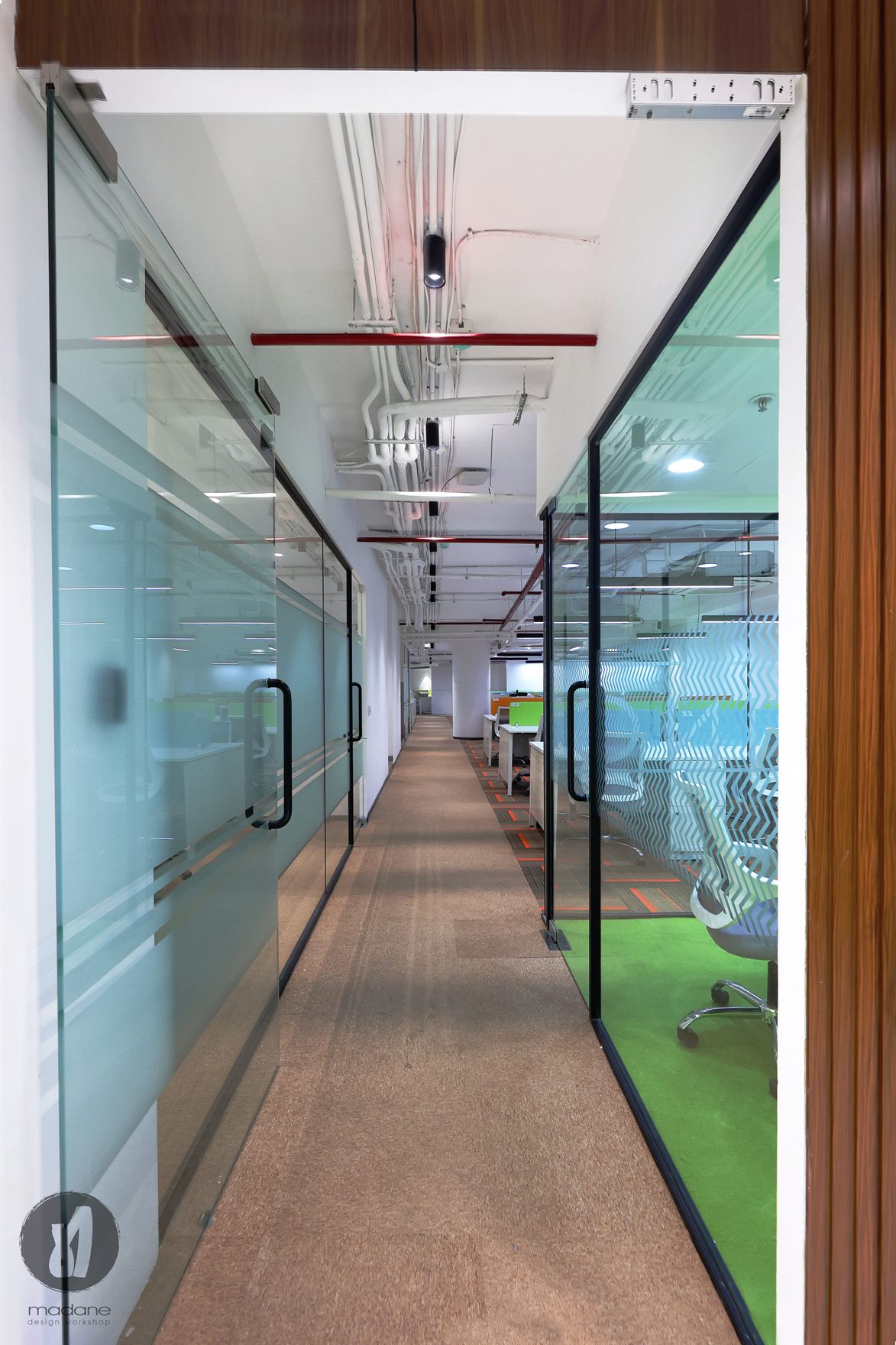 Glass-partitioned passage with frosted chevron film revealing a green-carpeted cabin, under exposed red-piped services
