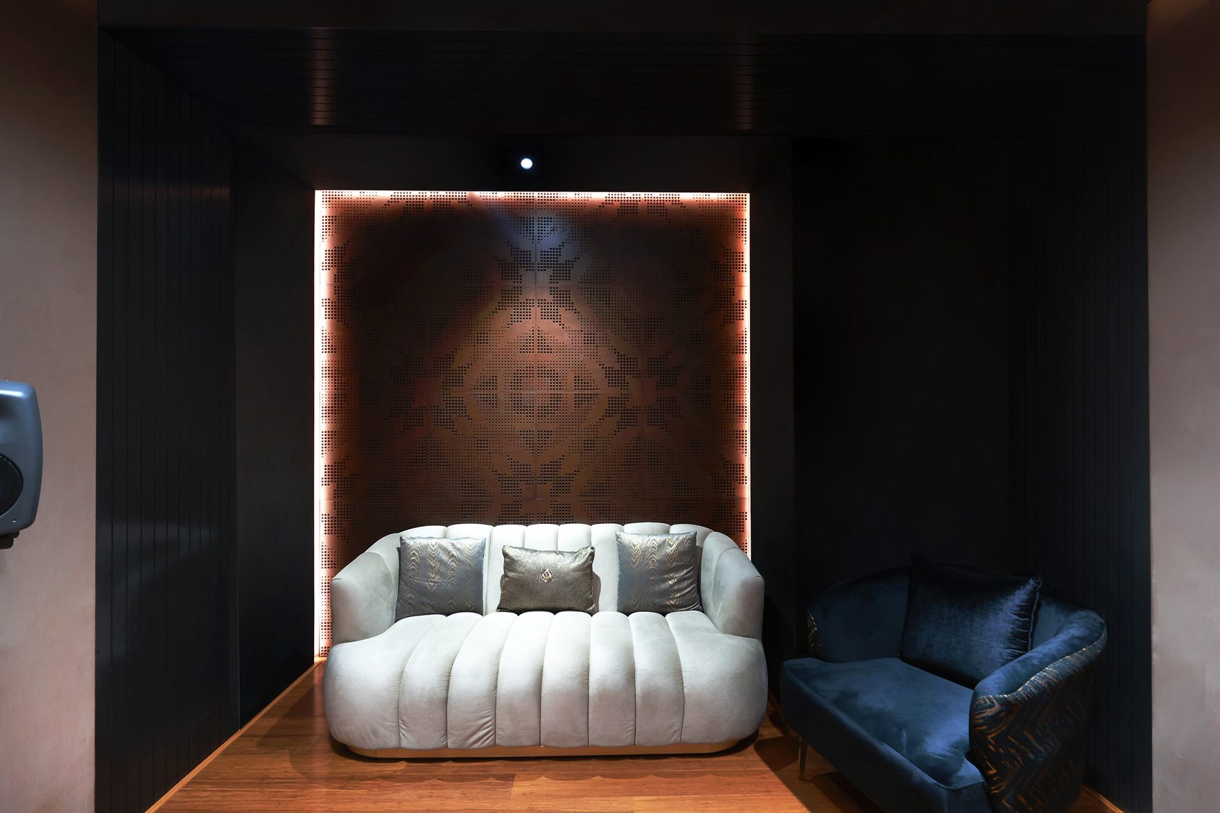 Lounge nook with a cream channel-tufted sofa and blue velvet armchair set against a backlit perforated-copper panel framed in black slatted walls over a wood floor