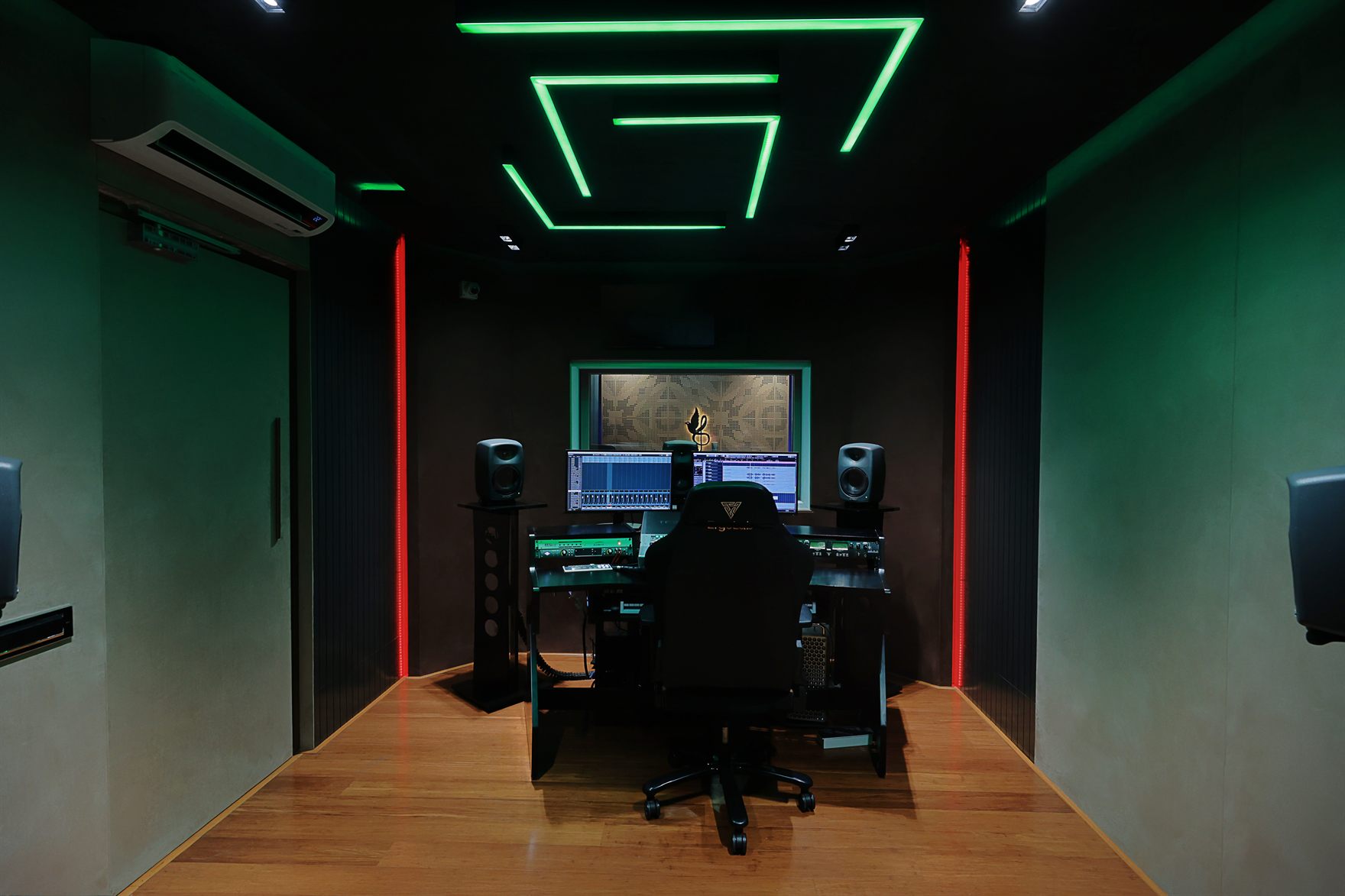 Control room with green neon geometric ceiling lines and red LED strips, studio monitors and a mixing desk of dual DAW screens facing a window into the booth