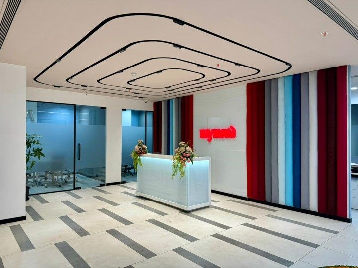 Reception with vibrant vertical multi-colour striped feature wall, red neon brand logo, illuminated front desk and concentric linear ceiling profile.