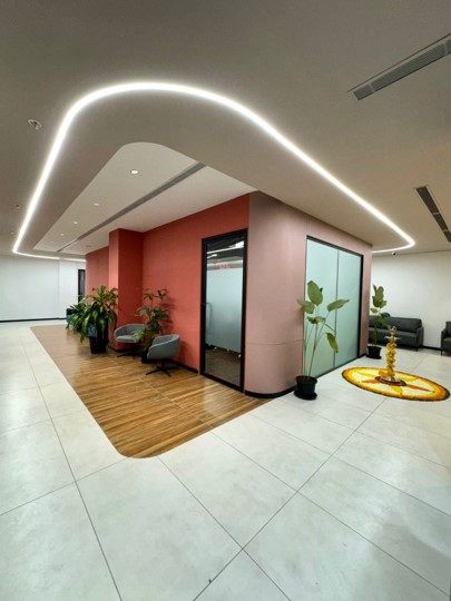 Breakout lounge with terracotta-toned glass cabin, wood-floor inset seating zone, planters and a curved floating ceiling with cove lighting.