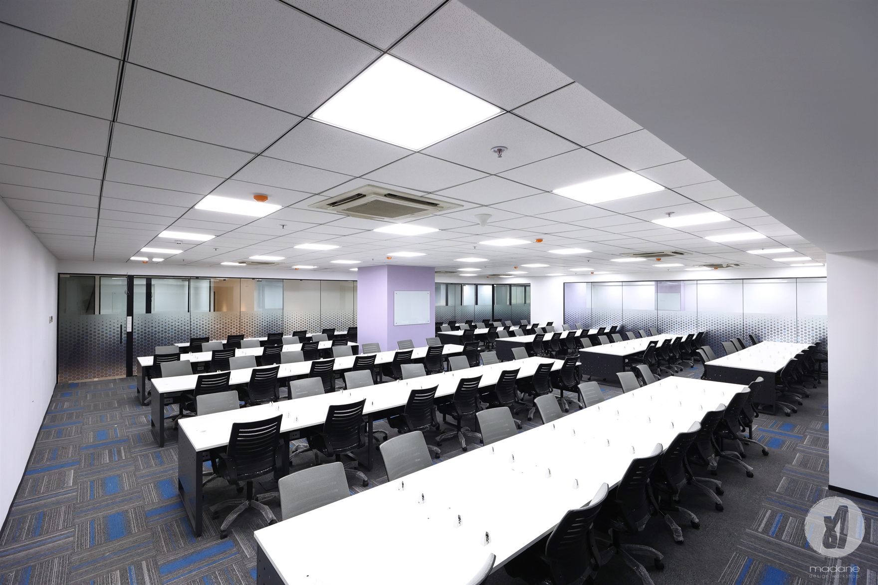 Large training hall with long rows of white desks and black mesh chairs under a grid ceiling, glass cabins with hexagon film along the walls