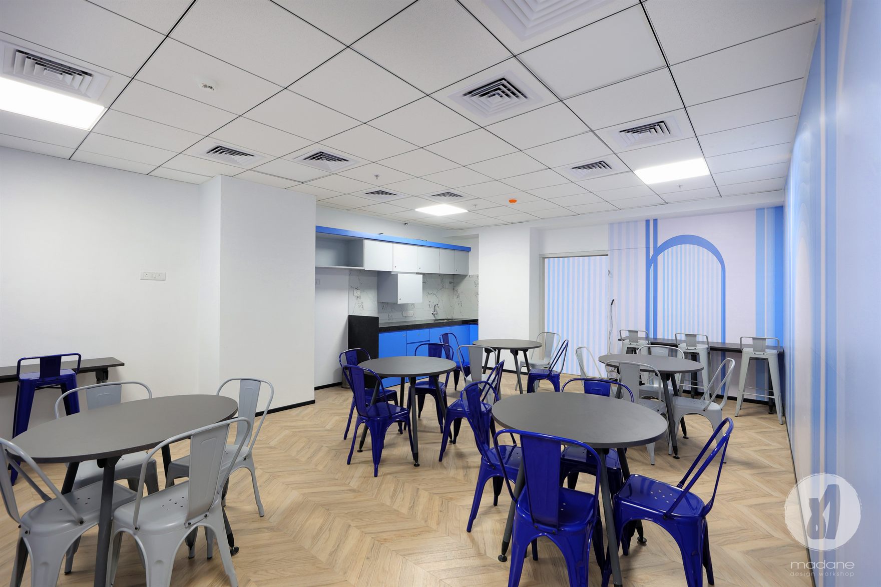 Cafeteria with cobalt and grey metal chairs around round tables, a blue pantry counter and an arch-motif wall on herringbone timber flooring