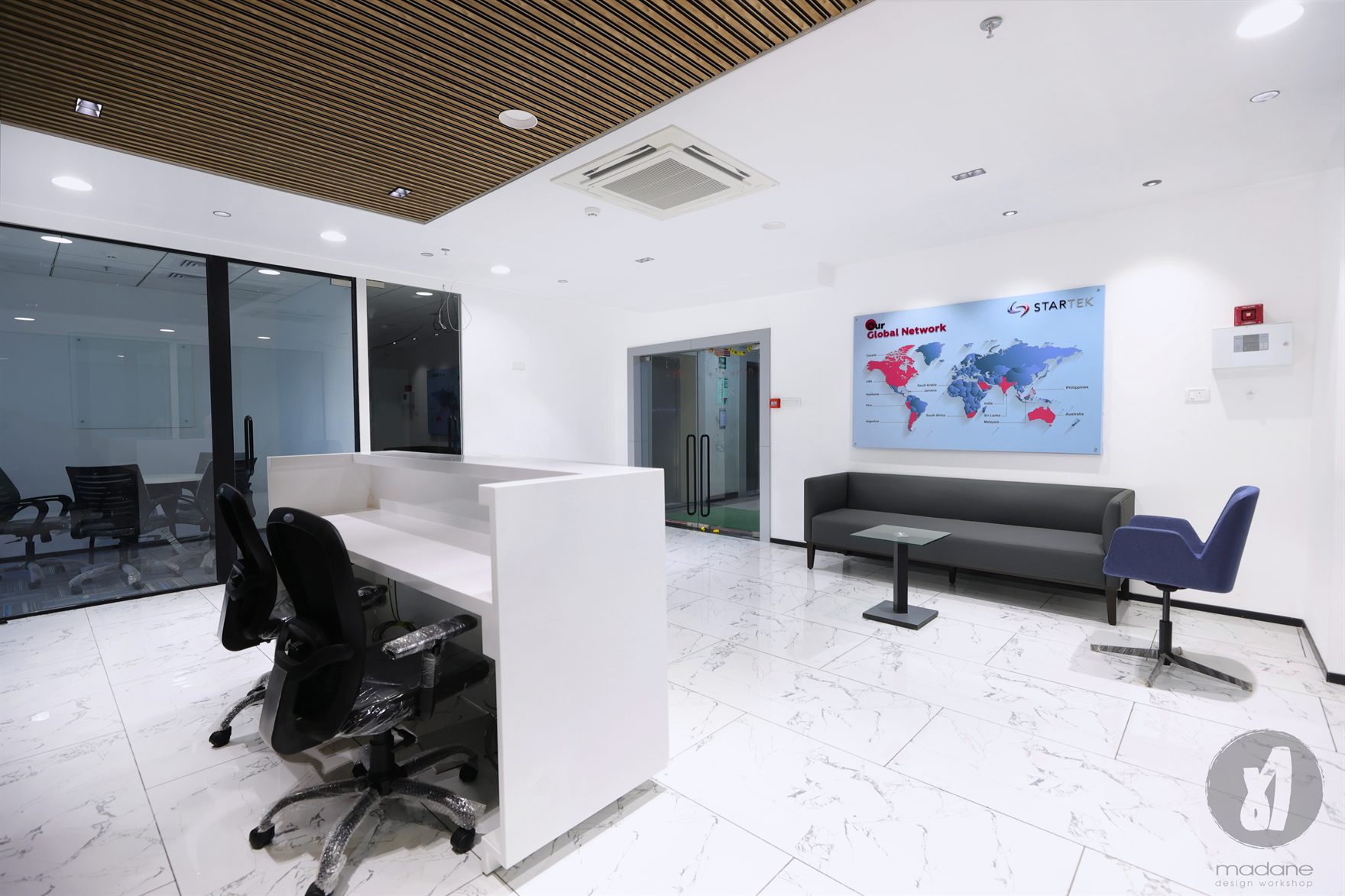 Reception with a white desk, timber-slat ceiling and a Startek Global Network world map, a grey sofa and blue tub chair on marble floor