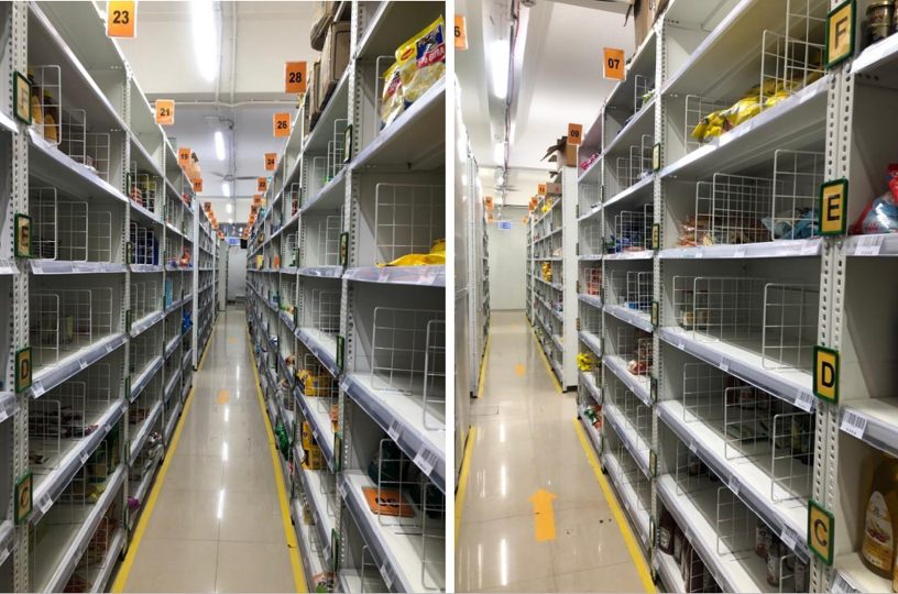 Dark-store aisles of white wire-mesh racking tagged with orange bay numbers and green zone letters, guided by yellow floor lines under bright linear lighting